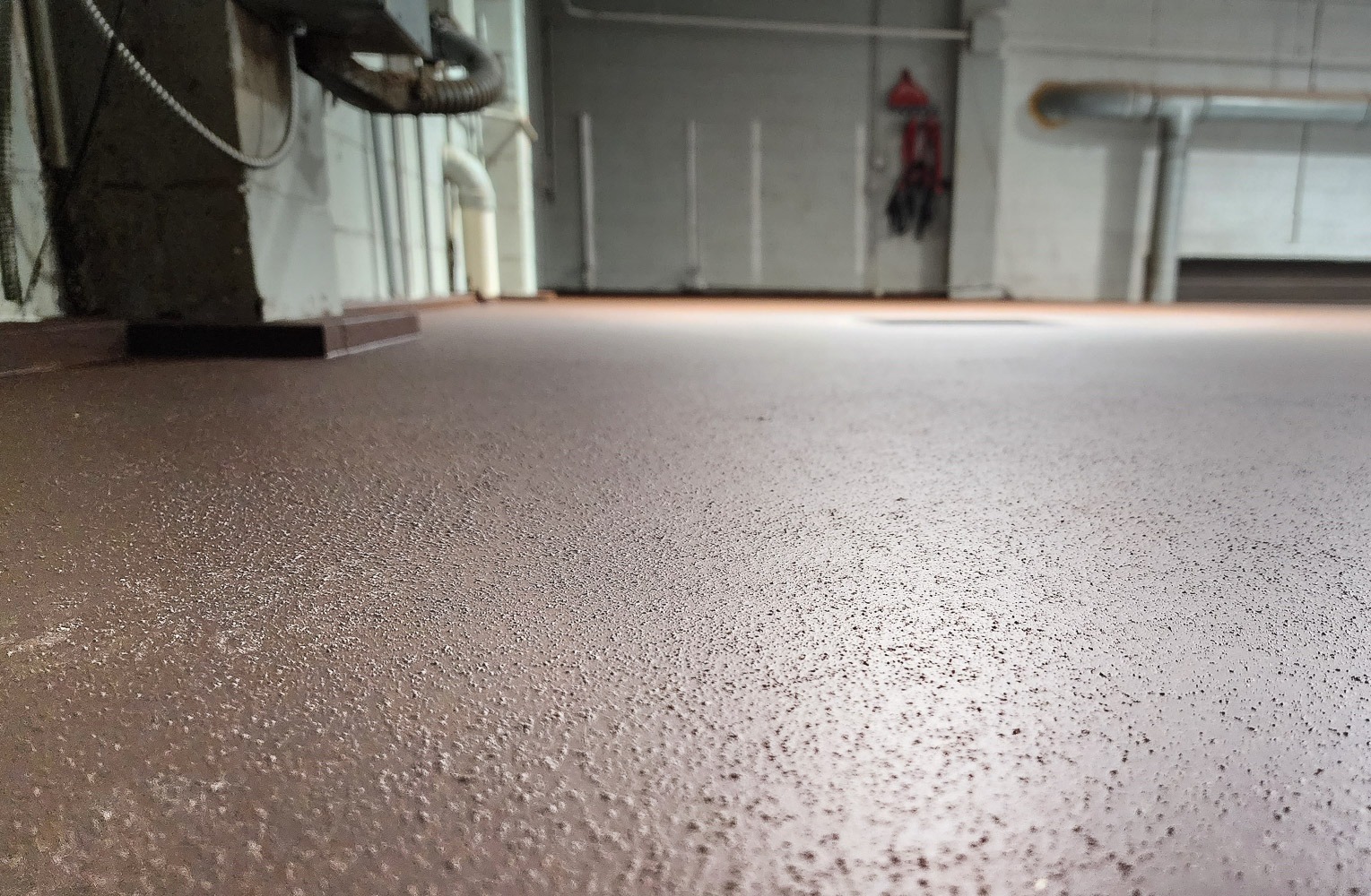 Close-up of brown AquaSeal UltraCoat non-porous thermoplastic coating system applied to an indoor facility floor, showing textured, slip-resistant surface near utility equipment