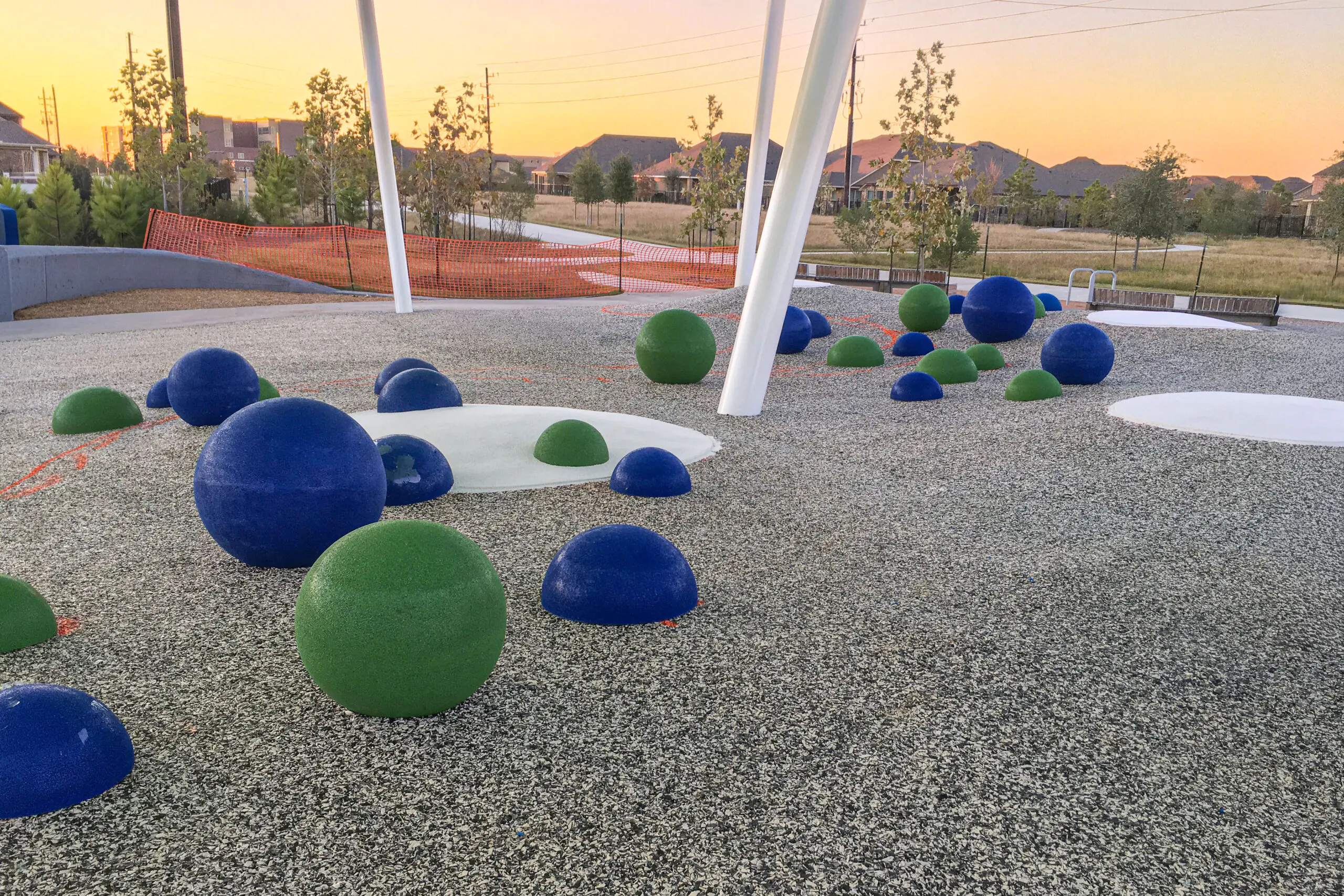 Green and blue AquaSeal EPDM rubber play spheres on a grey safety surfacing area at a modern playground during sunset, with shade structure posts and neighborhood houses in the background