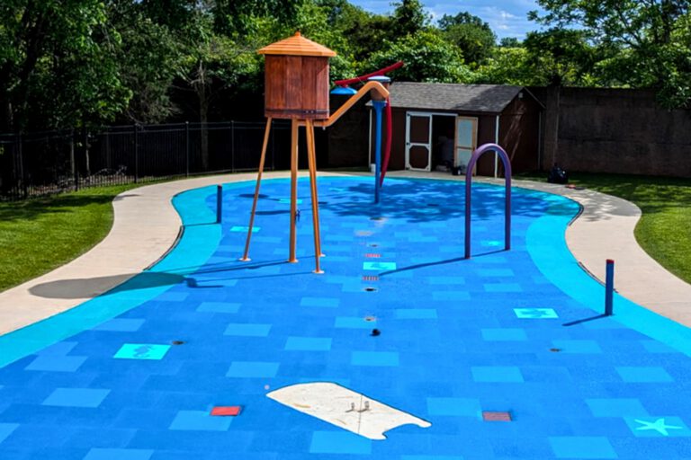 Wide view of a Aqua Tile resurfaced by AquaSeal Resurfacing LLC with blue patterned safety surfacing, a tipping bucket tower, spray features, and curved concrete walkways.
