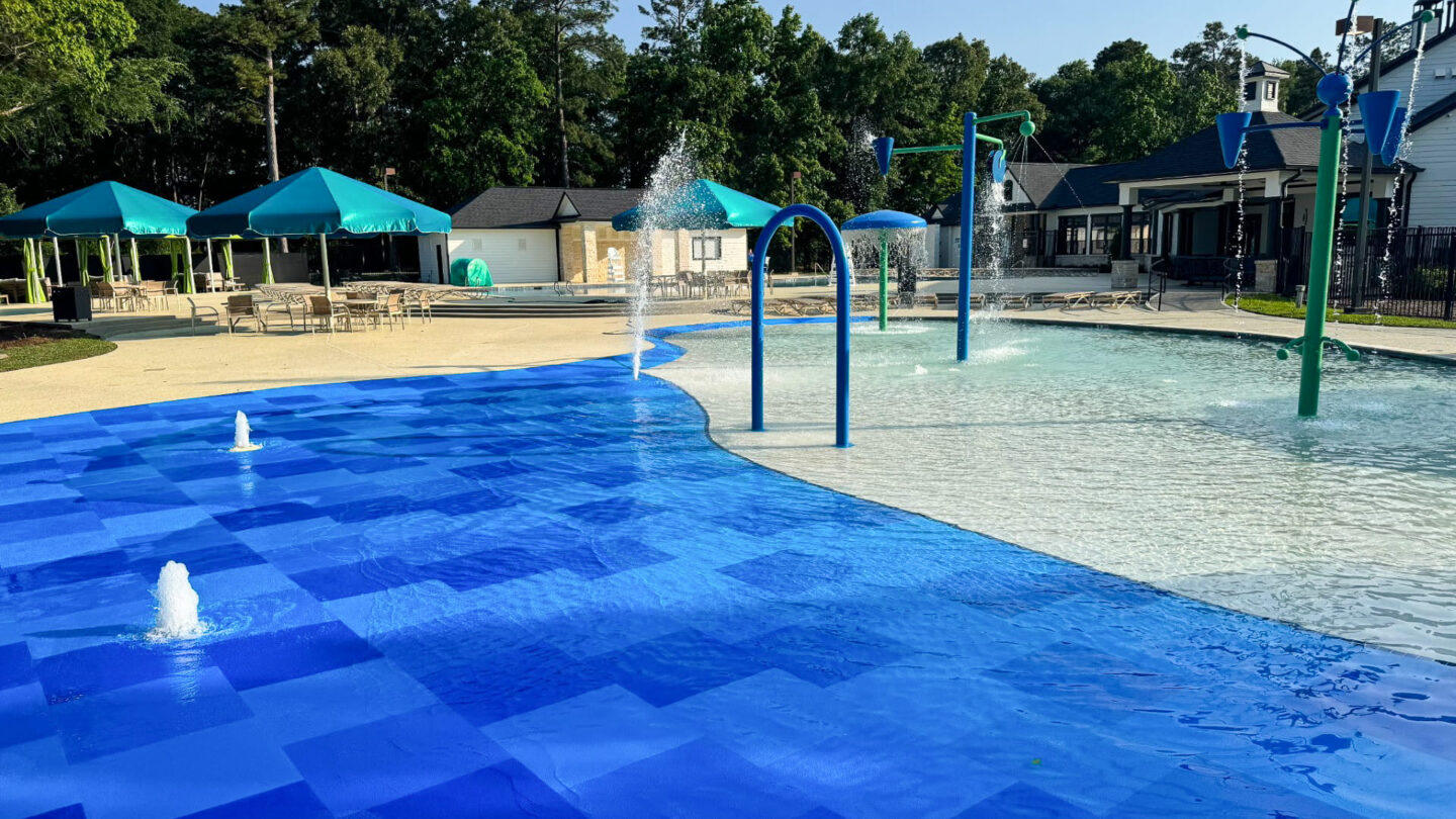 Blue splash pad safety surfacing installed by AquaSeal Resurfacing LLC next to a shallow pool, with spray jets, arched water features, and shaded seating in the background.